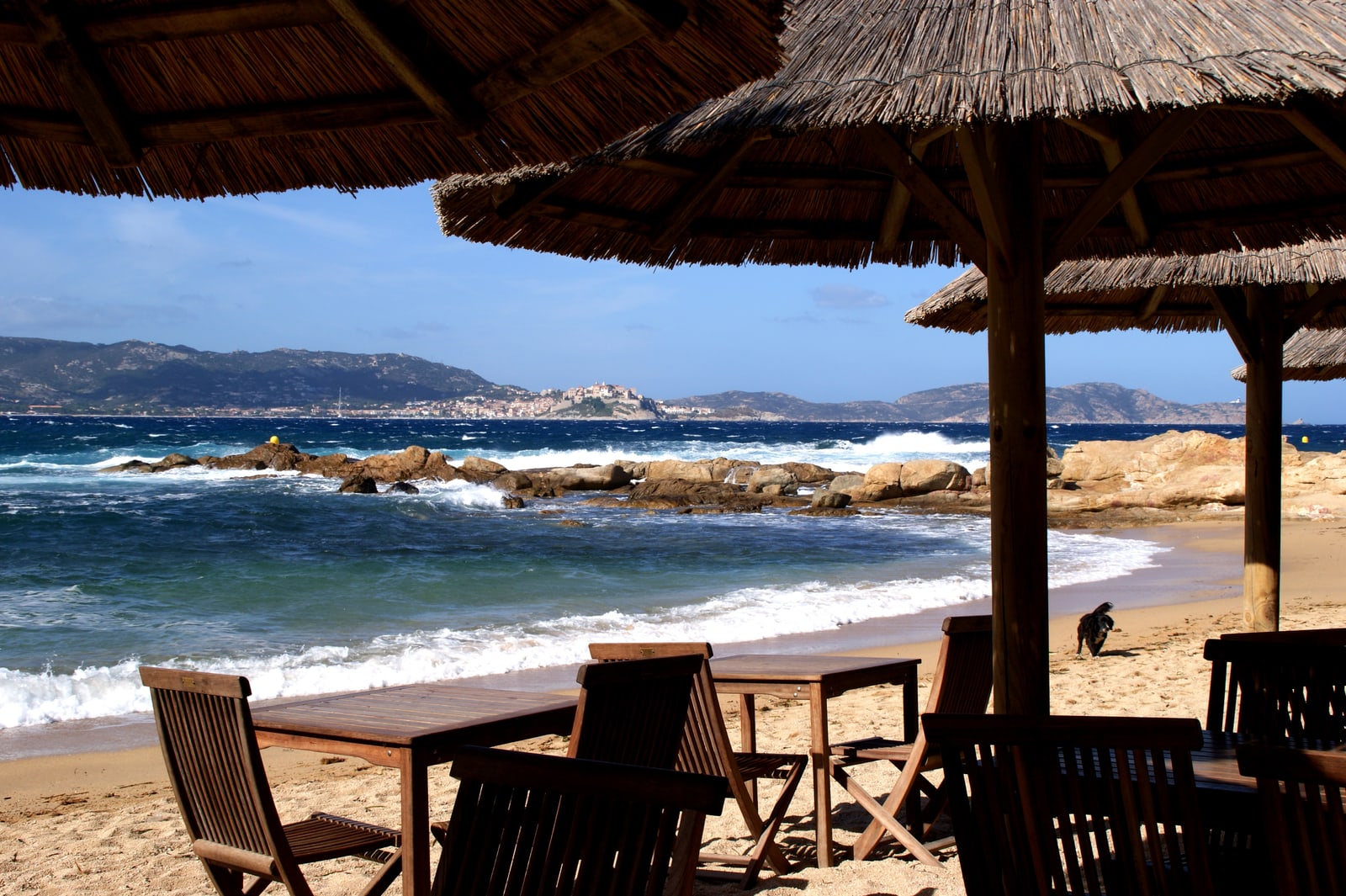 Beach bar with thatched parasols and wooden tables on the sand, looking across the sea to Calvi's citadel